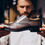 Homme mesurant son pied à la maison avec un papier de traçage et un mètre ruban, assis sur un sol en bois, vêtu de vêtements décontractés, lumière chaude et volumétrique du matin, atmosphère cosy et naturelle.