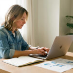 Une femme en reconversion professionnelle travaille sur un ordinateur, entourée d’un carnet ouvert et de graphiques marketing sans texte, dans un espace de travail lumineux.