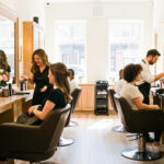 Intérieur lumineux et élégant d’un salon de coiffure, avec une coiffeuse accueillant une cliente et un coiffeur préparant son espace de travail.